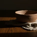 Tigela de cerâmica e pano de linho sobre mesa de madeira com luz dura lateral criando sombra alongada